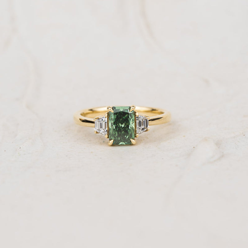 Studio shot of lab created emerald radiant cut diamond ring with green and clear stones laid flat with neutral textured background