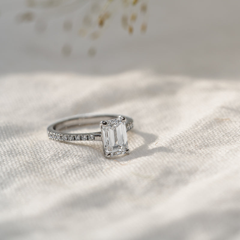 Platinum shoulder grain set emerald cut diamond thin band engagement ring laid flat on textured cloth material
