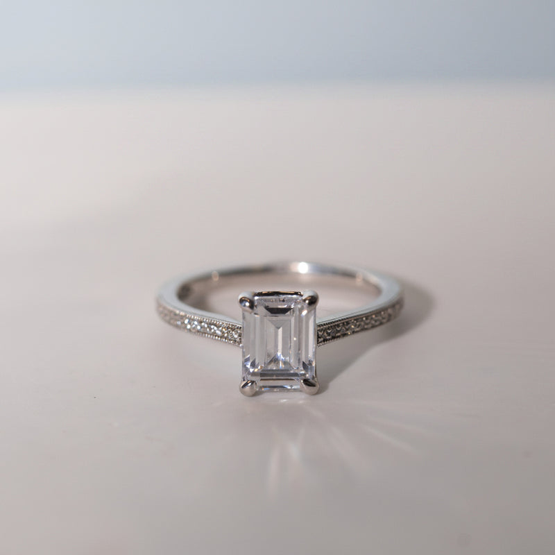 An emerald cut lab diamond shoulder set engagement ring with smaller diamonds set in a channel pattern around the main stone, displayed on a white surface with a blurred background.