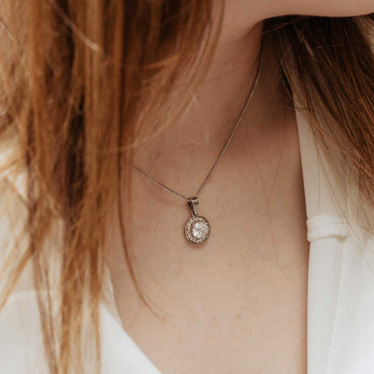 A person wearing a white shirt showcasing a round diamond halo pendant necklace with a halo of smaller diamonds, set in white gold.