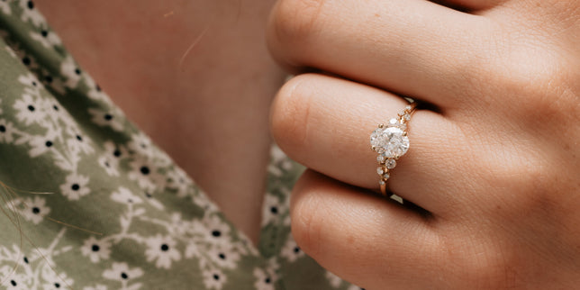 Accented engagement ring with oval cut centre in yellow gold is worn on woman's hand, her green dress can be seen in background.