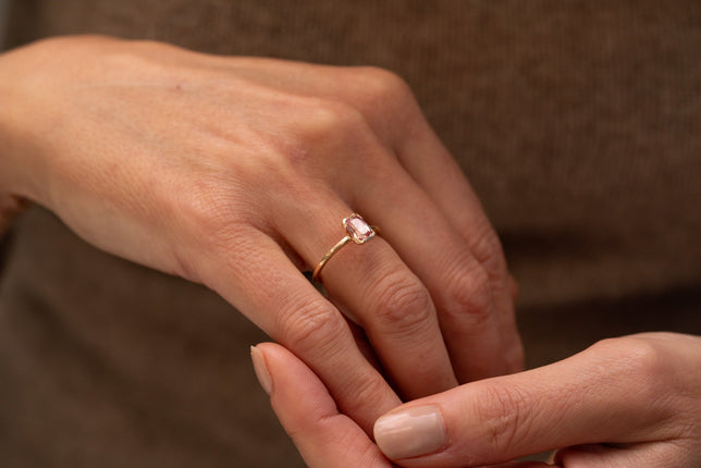 Close up of pink emerald cut engagement ring with channel set side stones, worn on model wearing a grey jumper. 