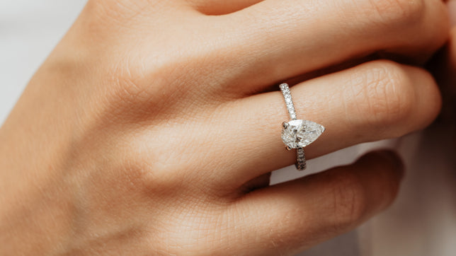 Close up of pear cut lab diamond engagement ring with lab diamond set band in Platinum worn on models hand. 