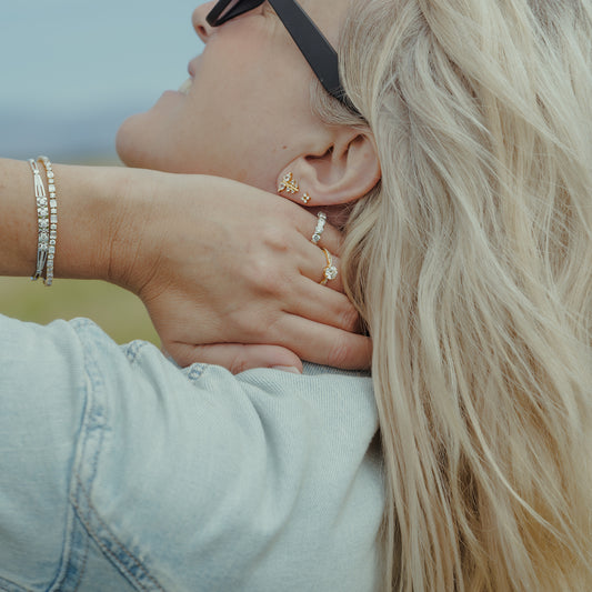 A close-up lifestyle shot of a woman with blonde hair wearing multiple delicate gold and silver earrings, including a butterfly earring, stacker earrings, and a bracelet, while wearing a light denim jacket.