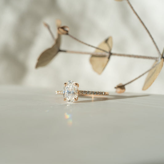 Rose gold ring with an circular oval shaped diamond on a light background and dried flower detail in midground