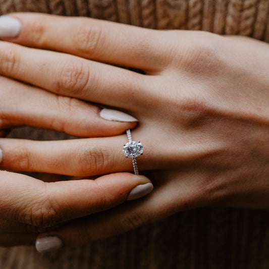 Model putting on a minimalist slim oval lab created diamond engagement ring with four rounded claws wearing rust coloured jumper