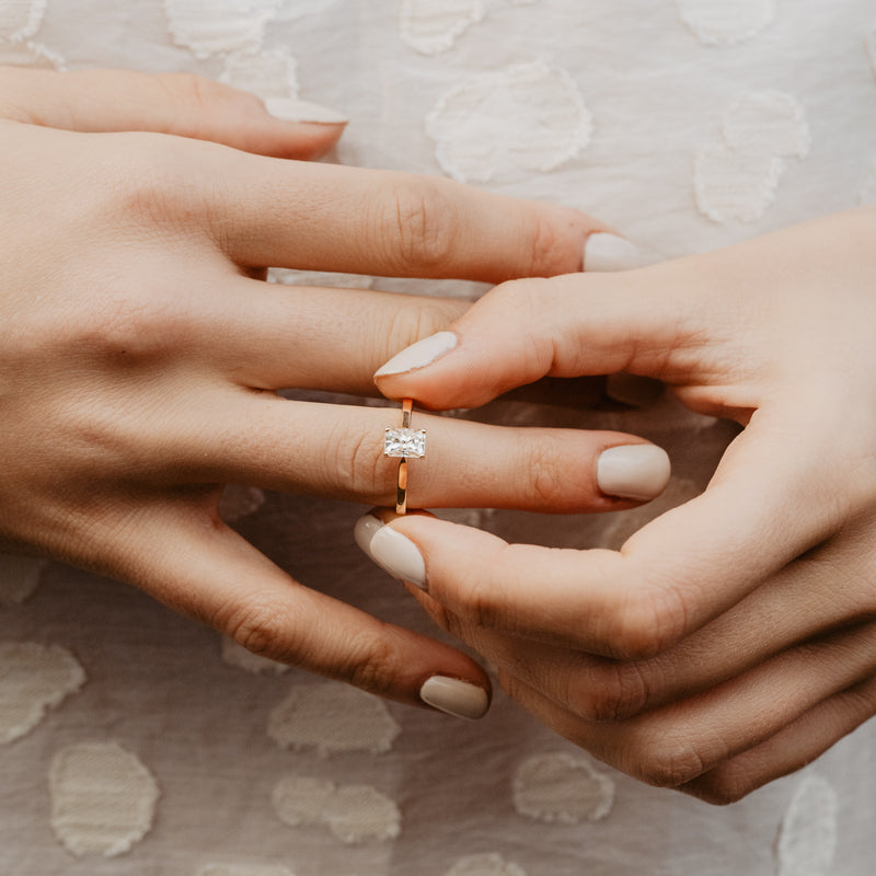 Close-up of two hands with an emerald cut diamond ring with a square flat profile thin band on a textured fabric background
