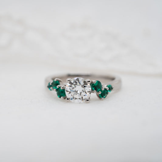 Flat lay of asymmetrical diamond engagement ring with emerald green side stones and round centre diamond 