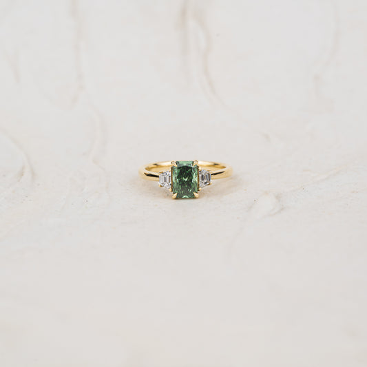 Studio shot of lab created emerald radiant cut diamond ring with green and clear stones laid flat with neutral textured background