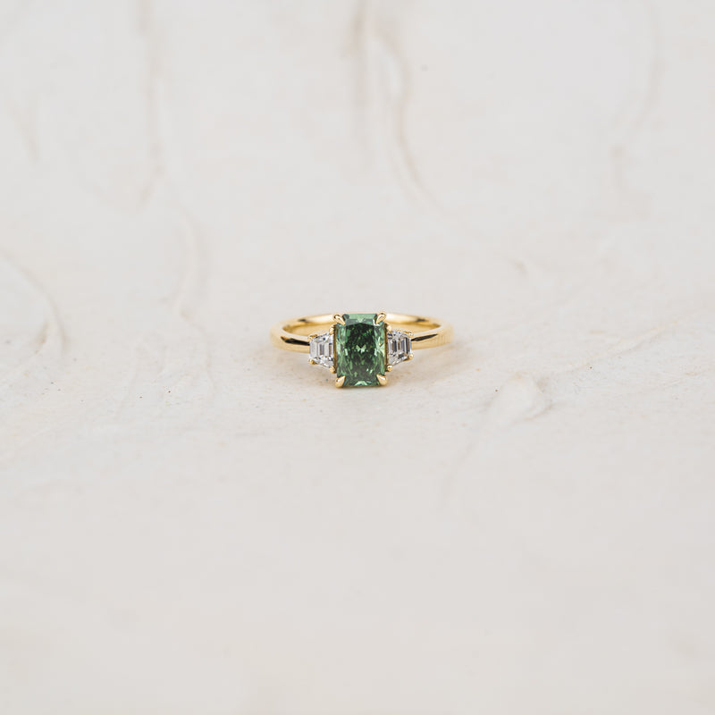 Studio shot of lab created emerald radiant cut diamond ring with green and clear stones laid flat with neutral textured background