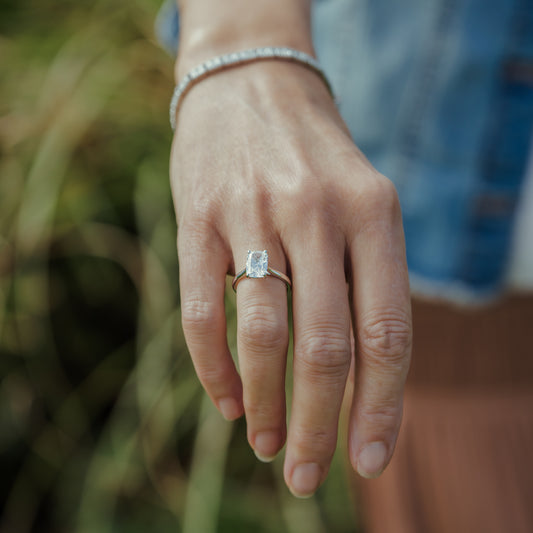 A close-up of a person's hand wearing a silver ring with a rectangular clear gemstone, with a matching silver bracelet on the wrist and a blurred outdoor background.