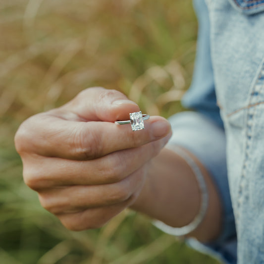 A close-up of a hand holding a platinum ring with an elongated diamond against a blurred outdoor background of dry grass.