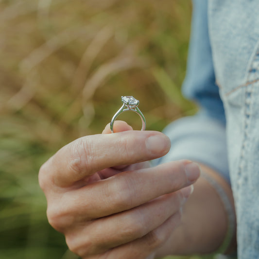 A platinum engagement ring with a cushion cut lab diamond held between fingers, with a soft-focus outdoor background of green and tan grass and a person wearing a light denim jacket.