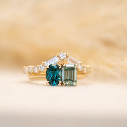 Gold ring with toi et moi green gemstones and diamond set baguette diamond wedding band on a soft beige background