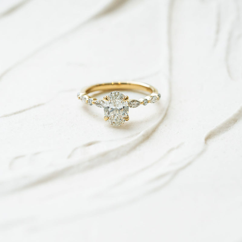 A yellow gold diamond ring with an oval-cut center stone and alternating small round and marquise shoulders, photographed on a softly textured white surface.
