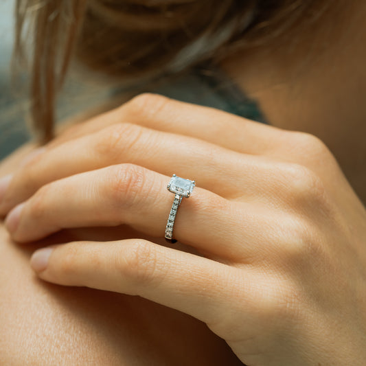 Model wears a platinum diamond ring with shoulder grain set pave diamonds running down shank and main diamond is held in by 4 claws 