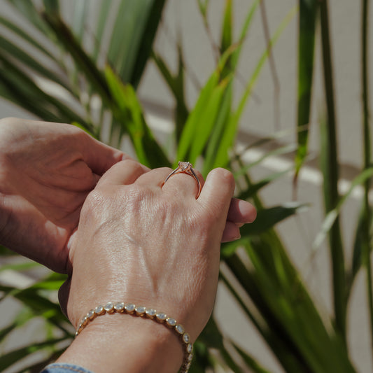 Close-up of hands with a princess solitaire engagement ring and gold tennis bracelet against a plant background