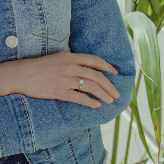 Person wearing a blue denim jacket with a rose gold diamond ring on their finger, standing in front of a plant.