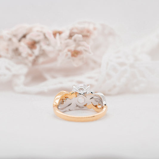 A large split tone 3 carat oval engagement ring with twisted split floral inspired band against a white blurred background