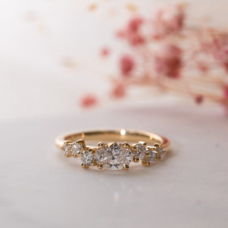 An oval lab diamond gold engagement ring featuring smaller lab diamonds set in asymmetric pattern with a floral pattern in the background.