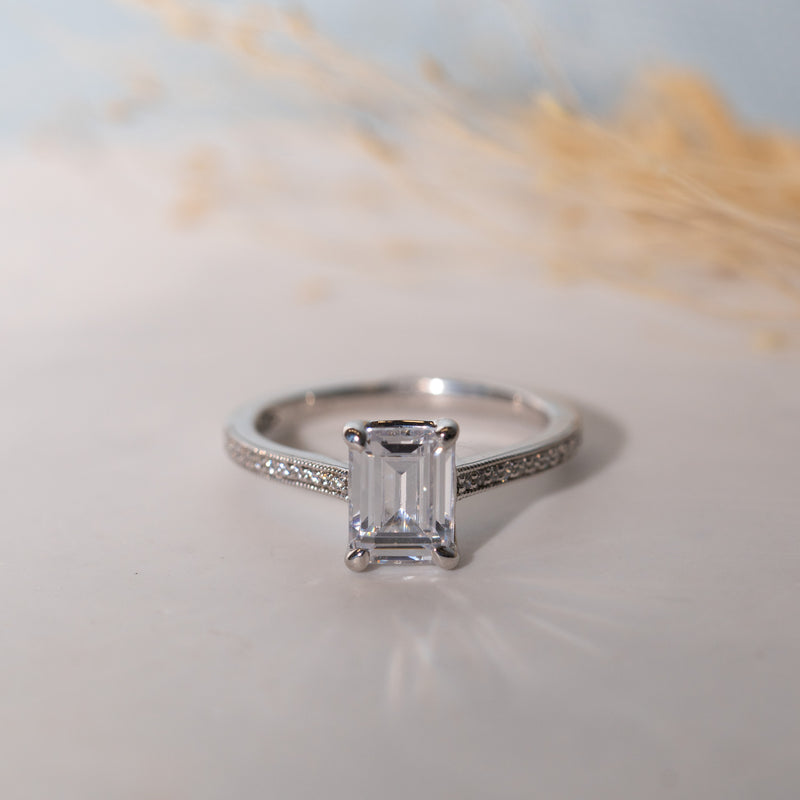 An emerald cut lab diamond shoulder set engagement ring with smaller diamonds set in a channel pattern around the main stone, displayed on a white surface with a blurred background.
