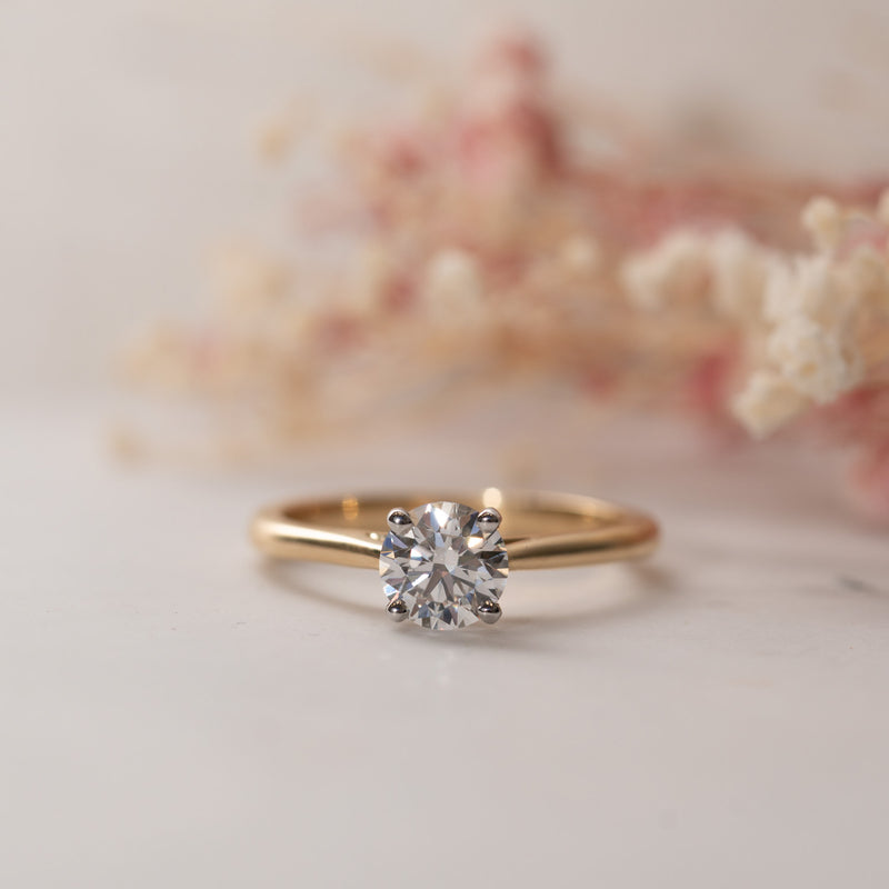 A round lab diamond classic solitaire engagement ring with a gold band, displayed against a floral background.