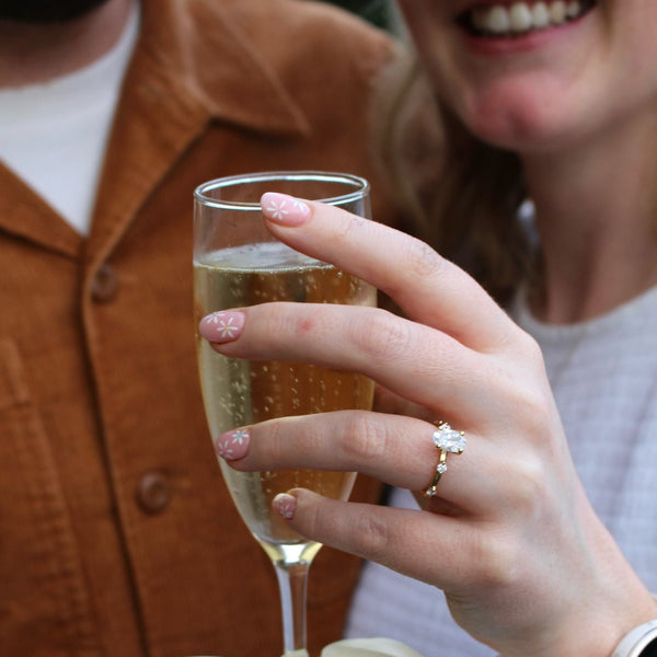 A close-up lifestyle shot of a person holding a champagne flute with a white satin bow tied to the base. The person is wearing a white striped shirt and a white smartwatch, and a gold ring with a clear gemstone is visible on their finger.