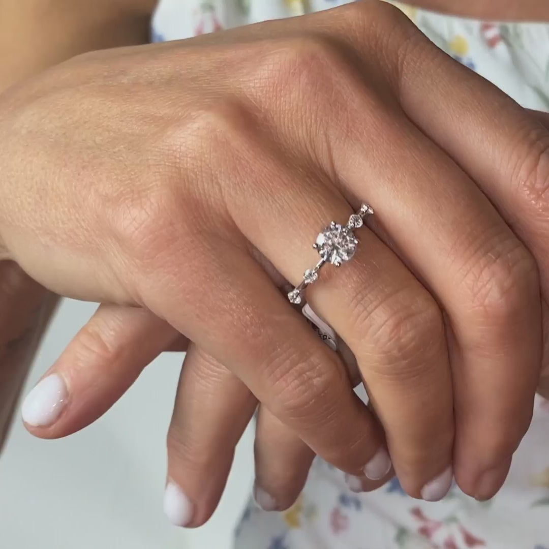 Model wears station ring with centre round diamond and accented shoulder orbiting diamonds and ultra thin band