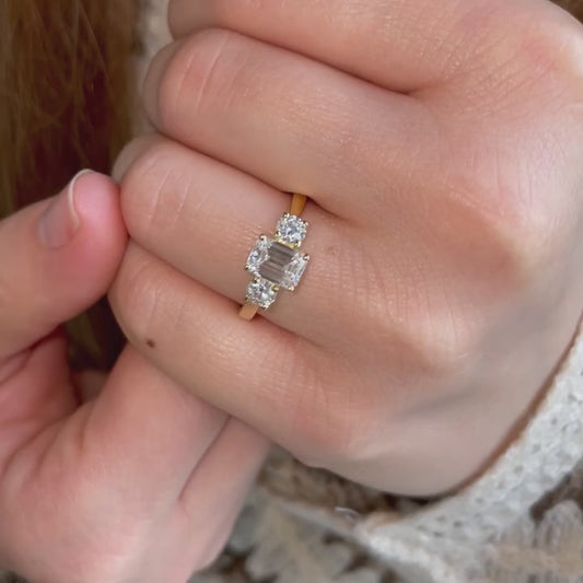 Model wears dainty emerald cut diamond ring in 18k yellow gold trilogy three stone design 