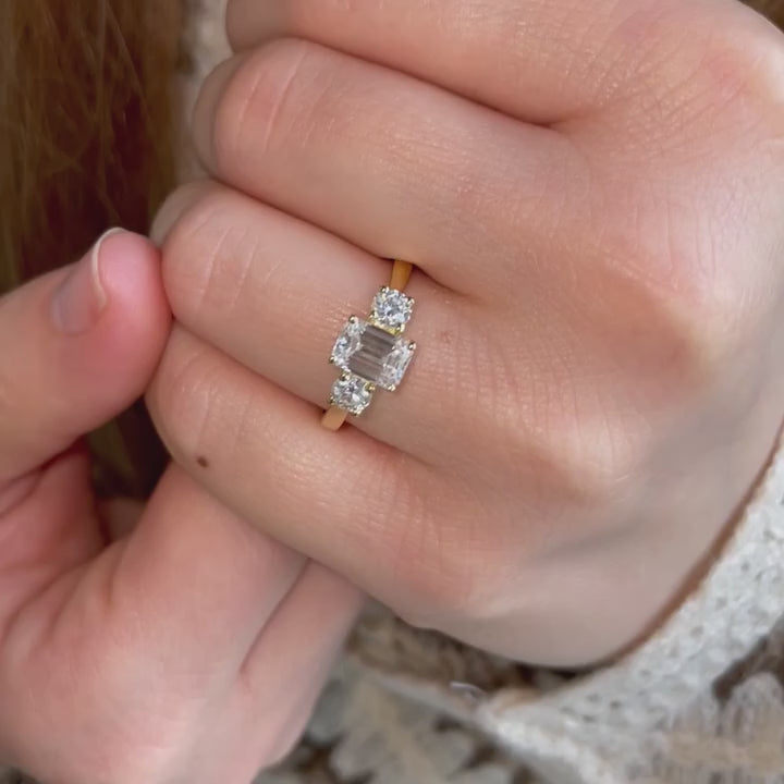 Model wears dainty emerald cut diamond ring in 18k yellow gold trilogy three stone design 